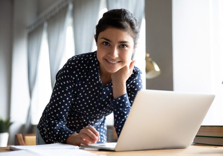 Portrait of smiling Indian girl standing at desk in living room working on laptop