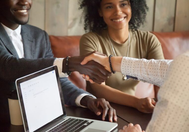 Happy young african american couple making deal handshaking caucasian insurance broker in cafe, black satisfied customer and realtor or sales person shaking hands at meeting in office with laptop